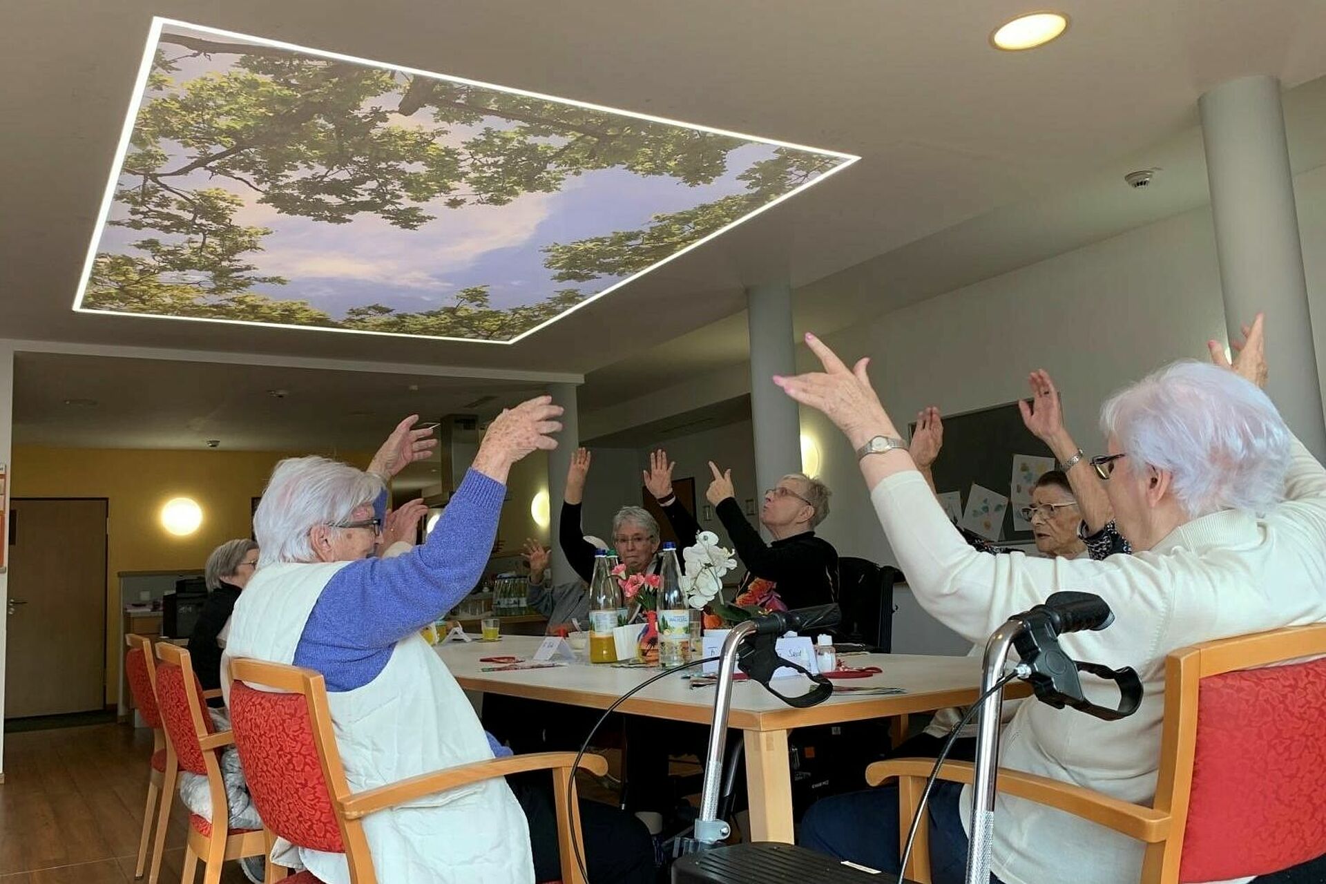 Leuchtdecke im Aufenthaltsraum. Darunter sitzen um einen Tisch Pflegeheim-Bewohner beieinander.